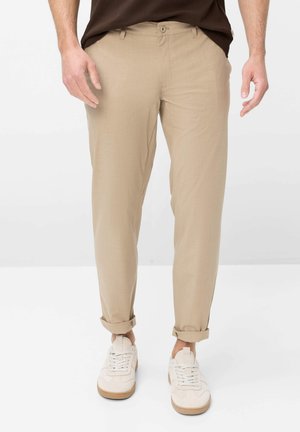 Homme portant un chino beige retroussé aux chevilles et des baskets blanches, debout devant un fond blanc uni.