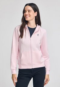 Felpa zip con cappuccio di colore rosa chiaro, realizzata in materiale morbido, dotata di tasca a marsupio, cappuccio con lacci e un piccolo logo nero sul petto.