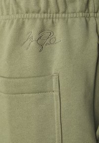 Pantalon de survêtement vert olive avec un tissu texturé, doté d'un détail signature brodé et d'une poche latérale. Taille élastique visible.