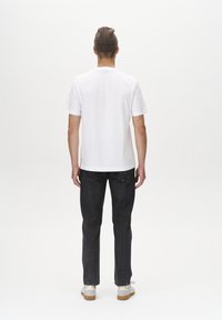 Camiseta blanca de manga corta, jeans índigo oscuros y zapatillas beiges. Lleva un gorro de punto marrón. Visto desde atrás contra un fondo liso.