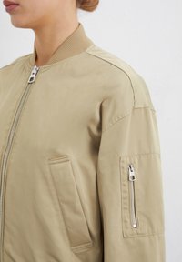 Veste bomber marron clair avec un col côtelé, des poches latérales et une poche à fermeture éclair sur la manche. Texture lisse et tissu léger.