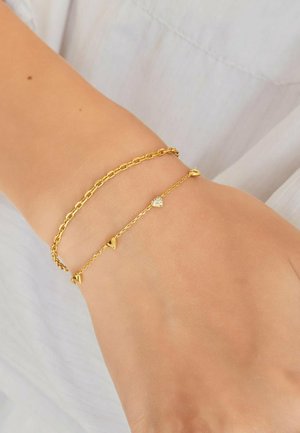 Handgelenk mit zwei zarten goldenen Armbändern, eines mit Kettenglieder-Design, das andere mit kleinen goldenen Herzen und einem zentralen Kristallherz-Anhänger.