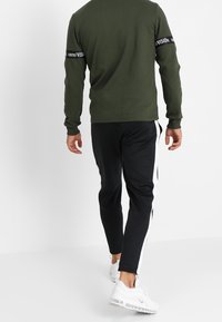 Completo da uomo con una maglietta a maniche lunghe verde oliva con scritte sulle maniche e pantaloni jogger neri a tapered con strisce bianche sui lati. Sneakers bianche.