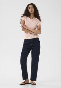 Hellrosa Kurzarm-T-Shirt, dunkelblaue gerade Jeans und schwarze Flip-Flops, stehend mit verschränkten Armen, neutraler Hintergrund.