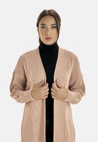 Ljust rosa stickad cardigan med ett texturerat mönster, öppen framsida och långa ärmar, lagd över en svart polotröja.
