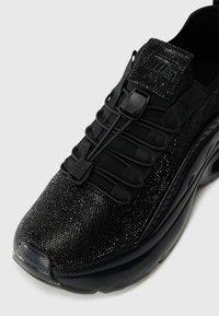 Chaussure de sport noire avec un bout lisse et brillant, un dessus texturé agrémenté d'accents scintillants, et un design sans lacets pour un enfilage facile.