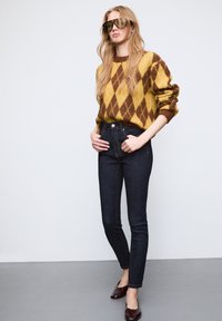 Sweter dziane w kolorze żółtym z brązowym wzorem w romby, ciemnoniebieskie dżinsy z wysokim stanem z przednimi kieszeniami oraz brązowe baleriny; modelka pozuje w okularach przeciwsłonecznych.