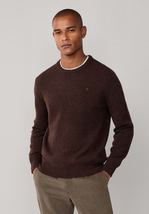 Mann mit kurzen Haaren, der einen dunkelbraunen Rundhalsausschnittpullover über einem weißen Hemd und beigen Hosen trägt, steht mit den Händen in den Taschen.