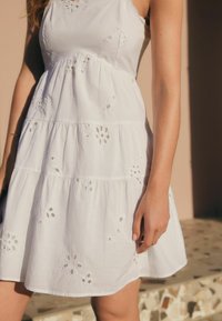 Robe en coton blanc avec des niveaux superposés, de la broderie florale et une conception sans manches. Texture douce et longueur au-dessus du genou.