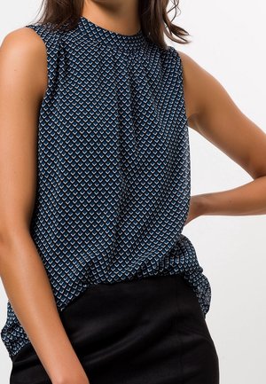 Blouse bleu marine sans manches avec petit motif géométrique blanc et bleu portée rentrée dans une jupe noire, bras plié à la taille.