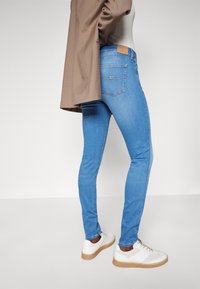 Jeans ajustados de mezclilla azul con cintura de talle medio, bolsillos traseros y una etiqueta de cuero en la pretina. Combinados con zapatillas blancas y un blazer marrón.