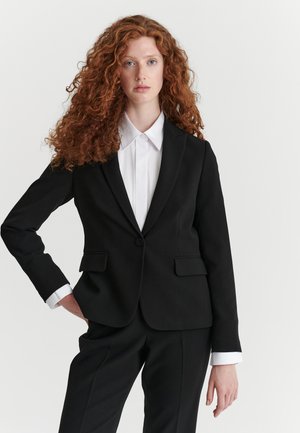 Femme aux longs cheveux roux bouclés, portant une veste de costume noire, un pantalon assorti et une chemise blanche, debout avec une main sur la hanche.