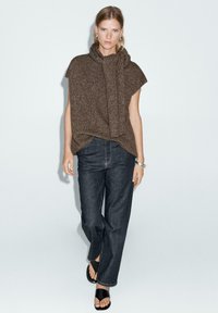 Poncho marrone in maglia testurizzata con colletto a cascata, abbinato a jeans blu scuro a gamba dritta e sandali neri con la zeppa. Bracciale in argento semplice come accent.