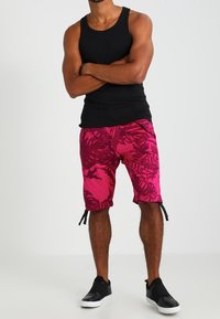 Rosa och svarta mönstrade badshorts med sidoknyten, med en tropisk bladdesign. Kombineras med en svart linne och svarta slip-on skor.