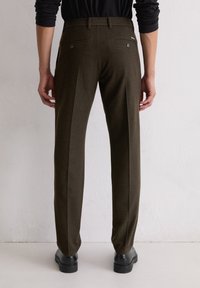 Slim-fit, donker olijfgroene broek met een gladde textuur, achterzakken en subtiele plooien. Gestoeld met zwarte schoenen.