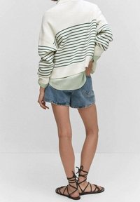 Person, die einen weißen Pullover mit grünen Streifen über einem grünen Hemd, blauen Jeans-Shorts und schwarzen, schnürbaren flachen Sandalen trägt, steht vor einer weißen Wand.