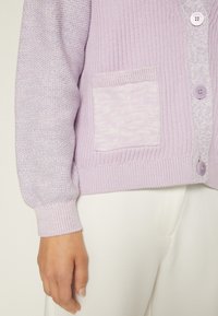 Lavendelfärgad cardigan med texturerad stickning på ena sidan och slät tyg på den andra. Har en ficka med lappar och matchande knappar.