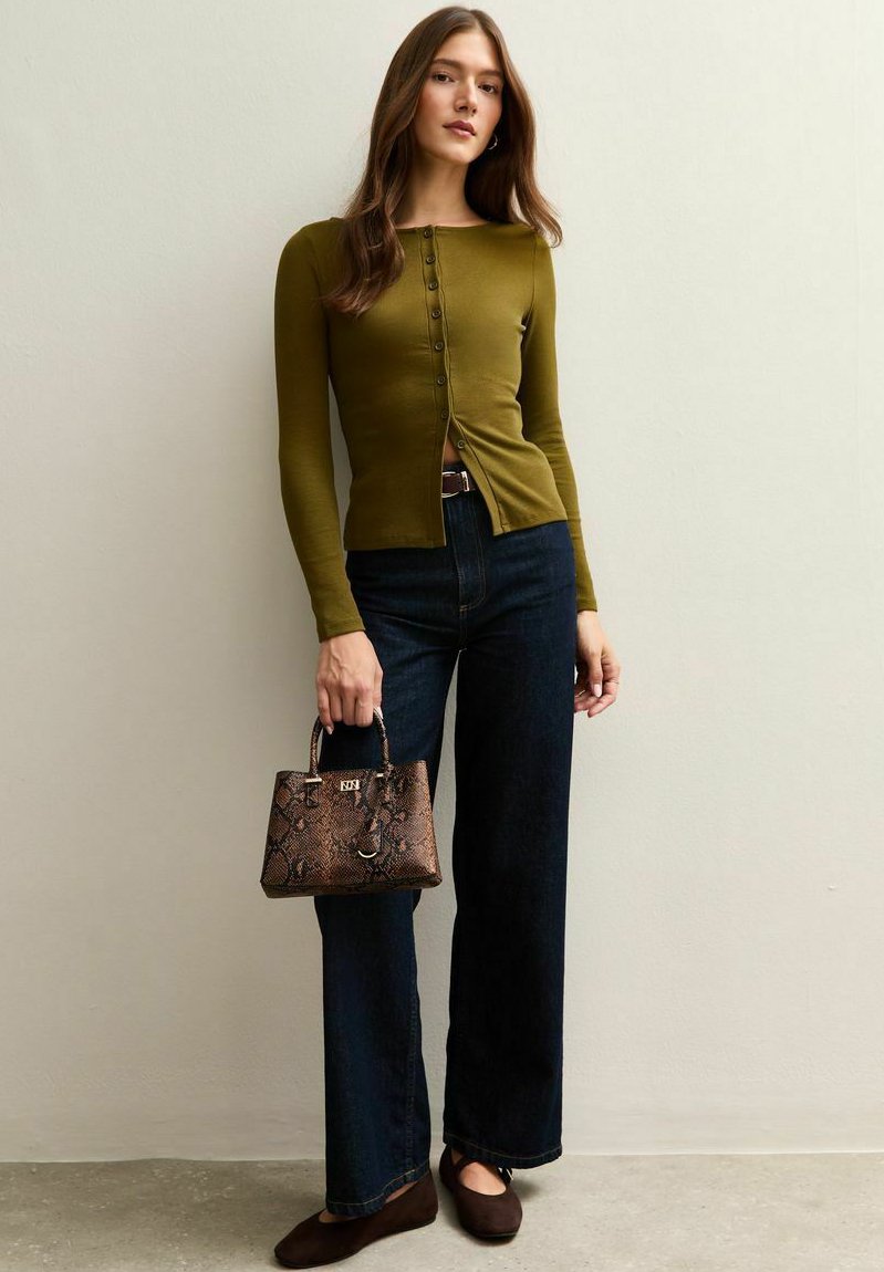 Dopasowany oliwkowozielony top z długim rękawem i guzikami, w połączeniu z ciemnoniebieskimi szerokimi spodniami jeansowymi. Trzyma brązową, teksturowaną torebkę z krótkim uchwytem.