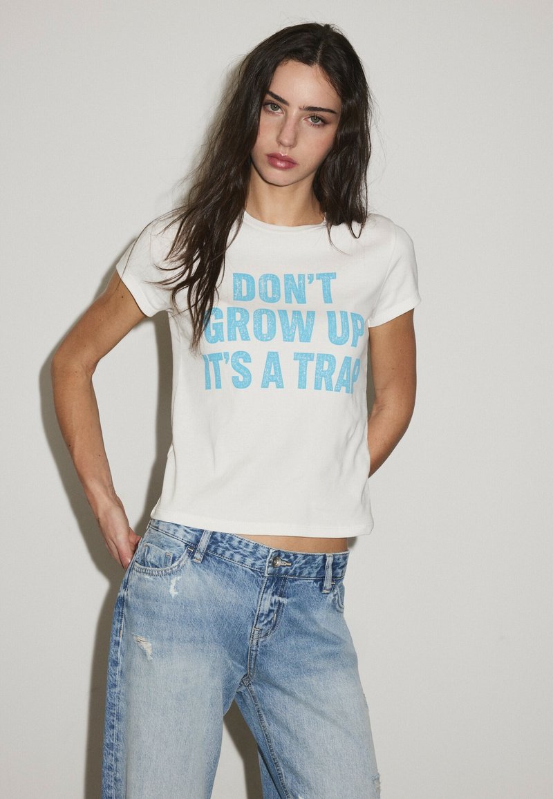 Woman with long dark hair wearing a white shirt with blue text "DON'T GROW UP IT'S A TRAP" and loose light blue jeans.