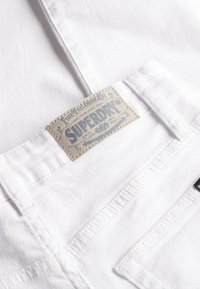Hvid denimstof med påsyet læderlap mærket "Superdry Fine Tailoring Co." nær lommens søm.