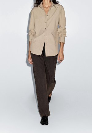 Woman wearing beige buttoned shirt with sleeves rolled up, dark brown wide-leg pants, and black shoes, standing against white background.