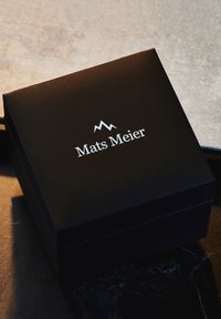 Schwarze rechteckige Box mit matter Oberfläche. Der Deckel zeigt den Namen "Mats Meier" in silberner Schrift und ein Berglogo darüber.