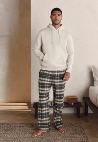 Hanorac gri deschis cu glugă, dotat cu buzunar frontal, asortat cu pantaloni de pijama cu carouri în nuanțe de navy, bej și verde. Modelul stă pe un covor țesut.