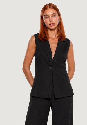 Donna con capelli rossi che indossa un blazer nero senza maniche e pantaloni neri abbinati, in piedi con le mani dietro la schiena su uno sfondo grigio chiaro.