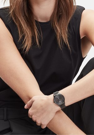 Femme en haut et pantalon noirs sans manches, assise les bras croisés, portant une montre bracelet métallique noire avec un cadran à motifs.