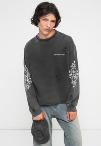 Joven con pelo rizado vistiendo una sudadera negra desgastada con diseños en las mangas en blanco y el texto "NO DESPERDICIES LA CULTURA", sosteniendo una gorra a juego.