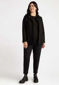 Chaqueta negra con capucha, cierre de botones y dos bolsillos frontales, combinada con pantalones negros y botas de tobillo. Las mangas acentuadas tienen pequeños remaches.