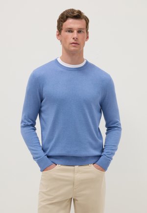 Giovane uomo con capelli castano chiaro, indossa un maglione azzurro chiaro sopra una camicia bianca e pantaloni beige, mani in tasca, sfondo neutro.
