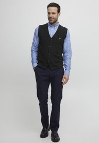 Svart ullväst över en ljusblå skjorta med knappar, bärs med marinblå byxor och svarta skor. Västen har en liten logotyp på bröstet.