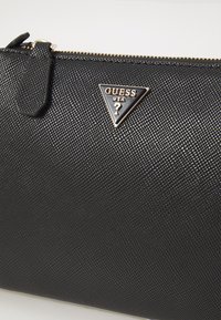 Juodas tekstūruotas piniginė su lygiu paviršiumi, su trikampiu metaliniu logotipu, kuriame nurodyta "GUESS USA" ir auksiniu užtrauktuko akcentu.