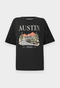 Juodas marškinėliai trumpomis rankovėmis ir laisvu siluetu, su grafiniu dizainu, vaizduojančiu kelią, kalnus ir kaktusus. Tekstas skaito "AUSTIN TEXAS."