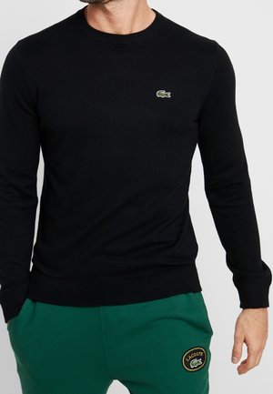 Mand iført en sort langærmet skjorte med en lille grøn krokodillelogo og grønne bukser med en Lacoste-patch på låret.