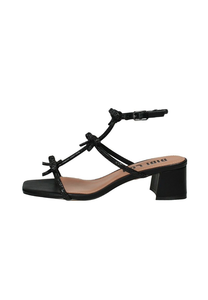 Sandalo nero con tacco, caratterizzato da una punta quadrata, sottili cinturini con dettagli a fiocco e un tacco grosso. Il materiale appare lucido con una texture sottile.