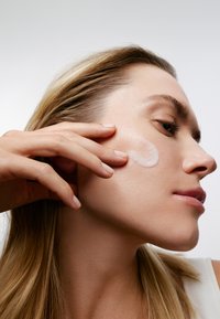 Crema hidratante aplicada sobre la piel; textura suave y blanca en una mejilla. La piel parece saludable y bien cuidada. Cabello de color claro.