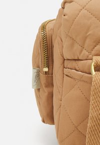Gequilteter, tanfarbener Rucksack mit goldfarbenem Reißverschluss und Stoffetikett. Seitentasche mit strukturiertem Detail und glatten Tragriemen.