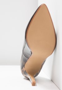 Chaussure à talon aiguille noire brillante à bout pointu montrée du dessous, avec une semelle beige et un petit talon carré sur une surface blanche.