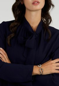 Blusa navy con colletto a fiocco. Il modello indossa due bracciali: uno in argento, di design intrecciato, e un bracciale nero elegante e lucido.