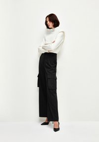 Weißer Pullover mit einzigartigem gepolstertem Schulterdesign, kombiniert mit schwarzen, gestreiften Cargo-Hosen und schwarzen spitzen High Heels, vor einem schlichten Hintergrund.