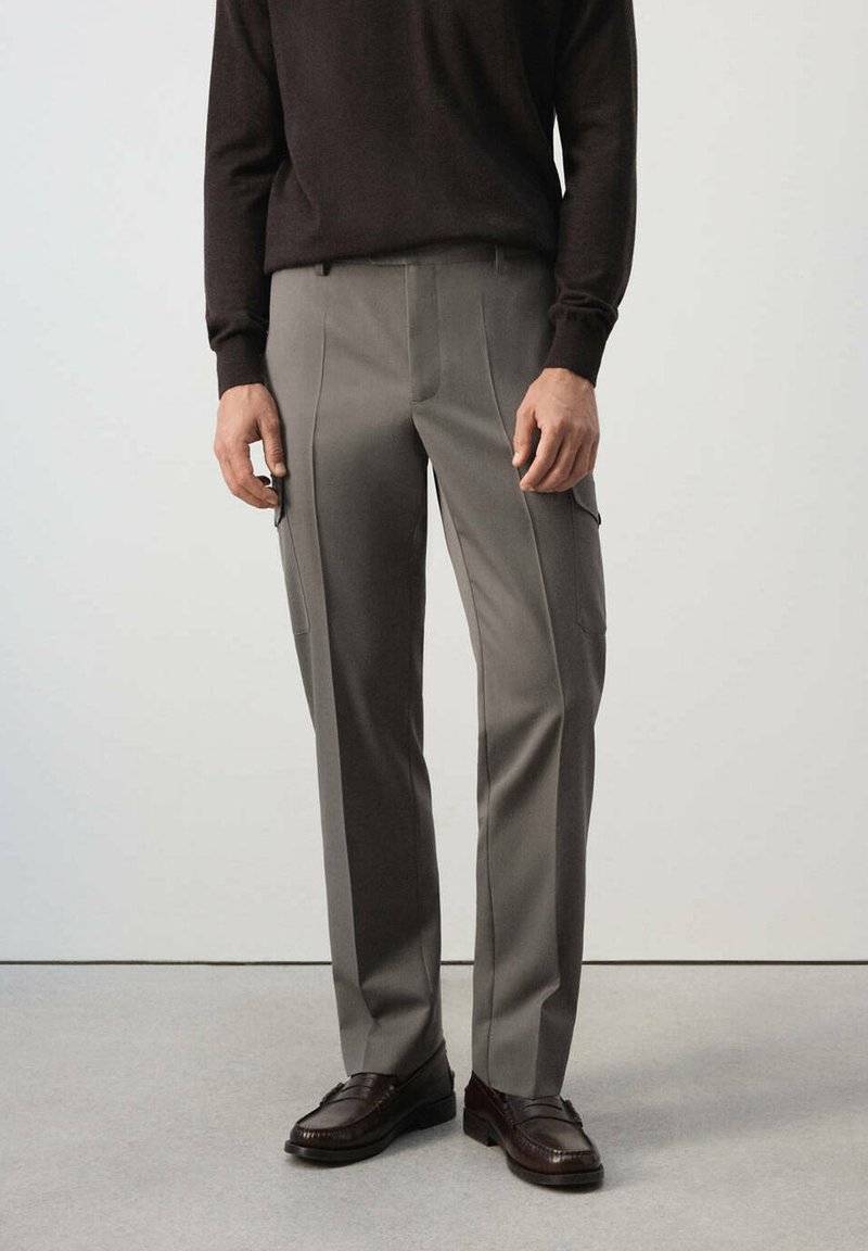 Homme portant un pantalon cargo gris à jambes droites, un pull marron foncé et des mocassins en cuir marron foncé, debout devant un fond uni.