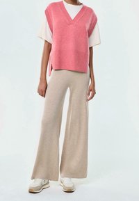 Pull sans manches rose tricoté avec un col en V, superposé sur un t-shirt blanc ; pantalon beige à jambes larges et baskets blanches avec des accents beiges.