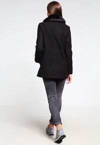 Abrigo negro con cuello de piel sintética, silueta ajustada y corte recto. Combinado con vaqueros grises y zapatillas de color gris claro, visto desde atrás.