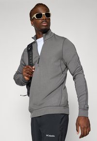 Pullover grigio con zip, realizzato in tessuto testurizzato, dotato di un collo mezza zip e dettagli delle cuciture in contrasto. Indossato con una camicia bianca sotto e uno zaino.