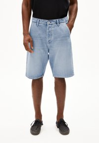Shorts en denim bleu clair avec une coupe droite, cinq poches, une taille mise en valeur et un ourlet net. Portés avec des baskets noires.