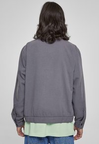Chaqueta gris con un corte relajado, con un dobladillo y puños de canalé, hecha de un tejido ligero, llevada sobre una camisa verde claro y unos jeans azules.