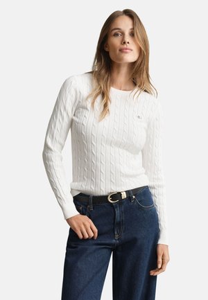 Hvid kabelstrikket sweater med lange ærmer, rund halsudskæring og diskret branding, kombineret med mørkeblå denimjeans og et sort bælte.