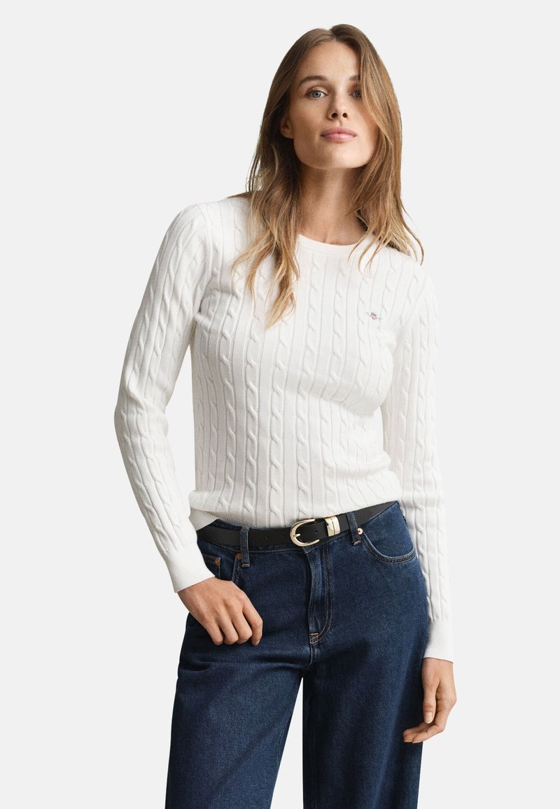 Weißer, kabelgestrickter Pullover mit langen Ärmeln, rundem Ausschnitt und dezentem Branding, kombiniert mit dunkelblauen Jeans und einem schwarzen Gürtel.